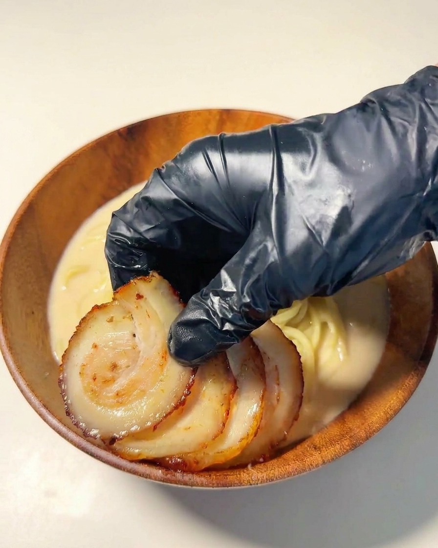 Una mano con guanto nero che dispone fette di chashu scottato sopra i noodles.
