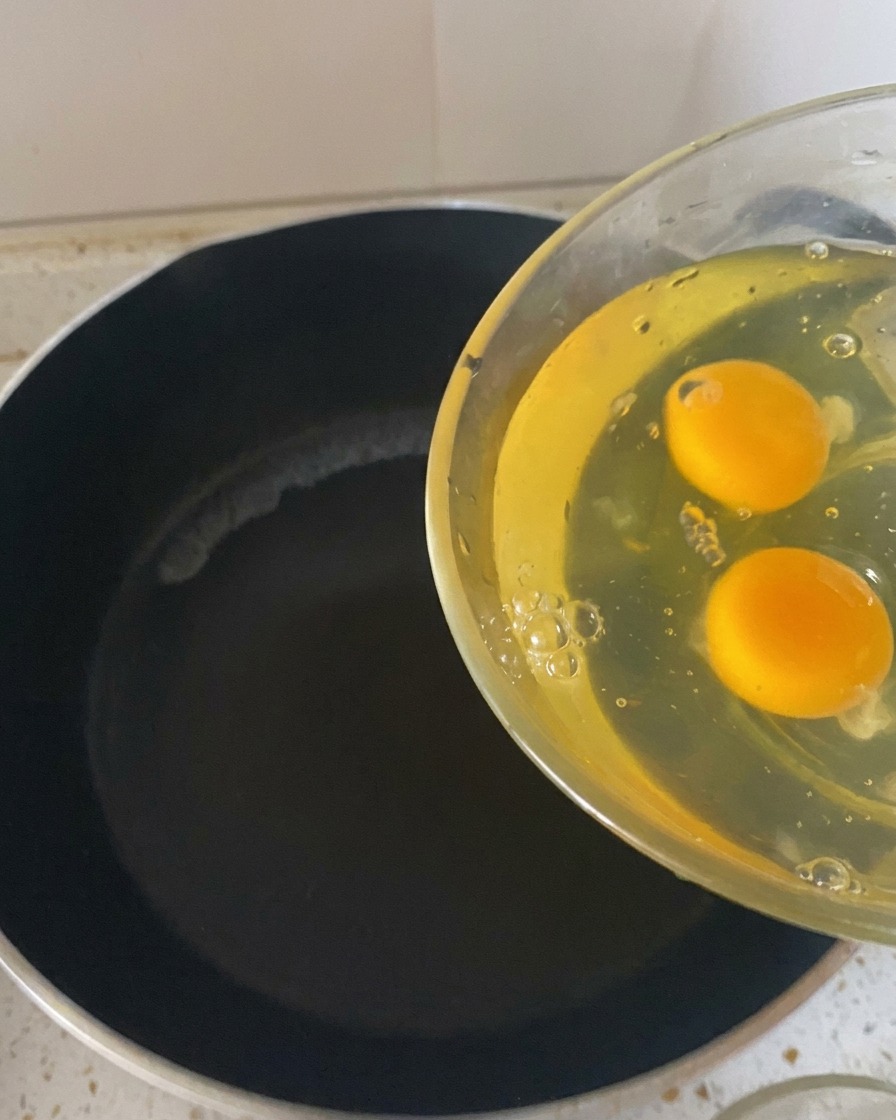 Due uova crude versate da una ciotola di vetro in una padella scura ben calda.