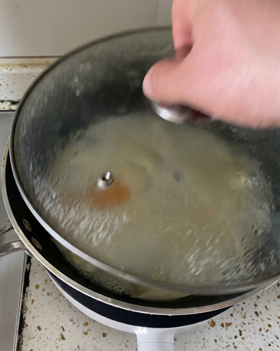 Una mano solleva il coperchio di vetro dalla padella per svelare vapore e uova cotte alla perfezione con formaggio fuso in superficie.