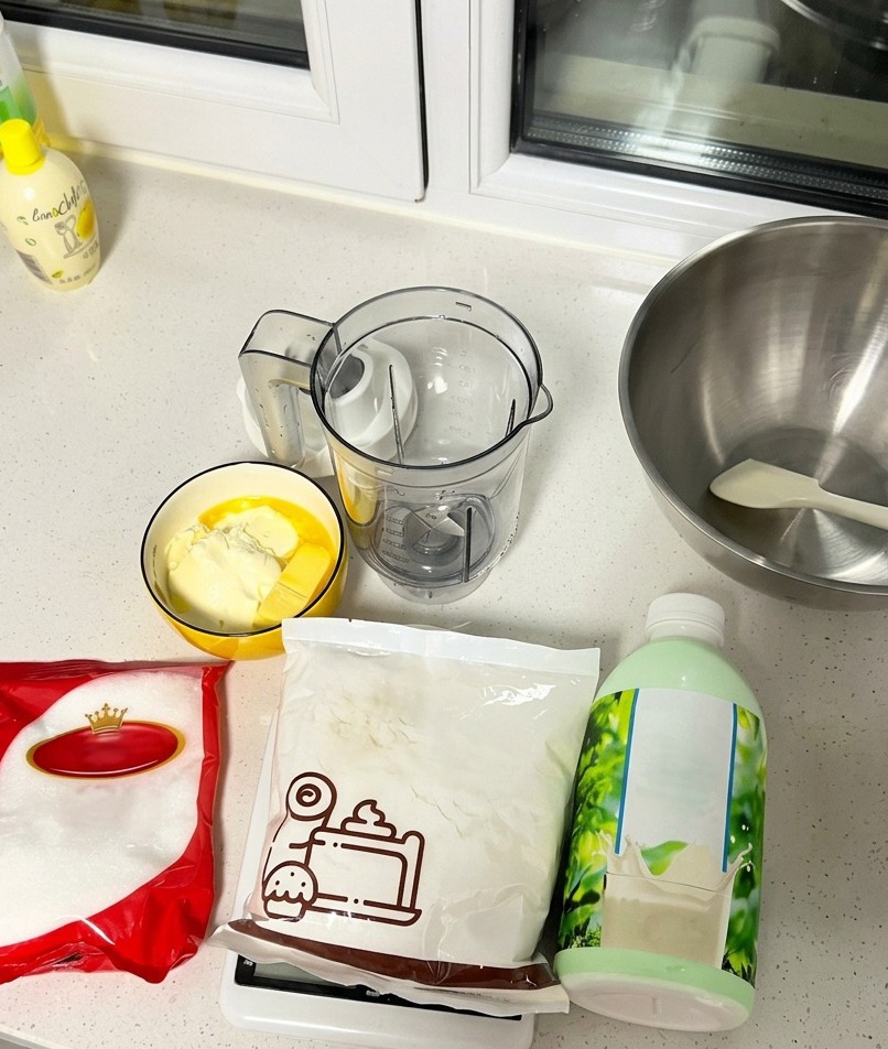 Ingredienti per la torta disposti sul piano da lavoro, tra cui una bottiglia di latte, zucchero, farina e una ciotola con burro e formaggio cremoso.