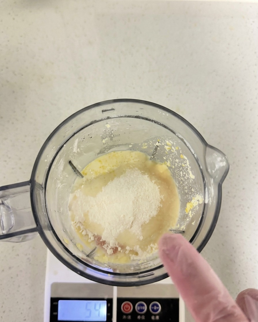 Farina e altri ingredienti secchi aggiunti sopra il composto umido allinterno del bicchiere del frullatore posizionato su una bilancia digitale.