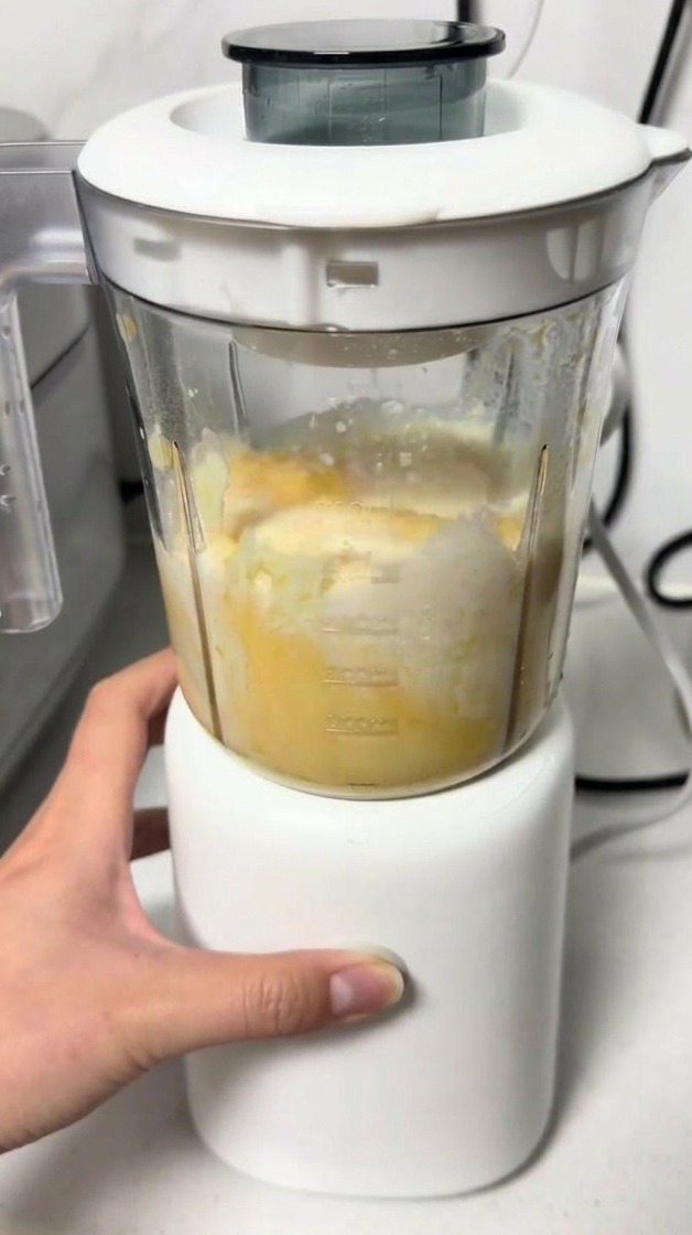 Una mano aziona un piccolo frullatore bianco per amalgamare limpasto giallo di formaggio e uova nel bicchiere trasparente.