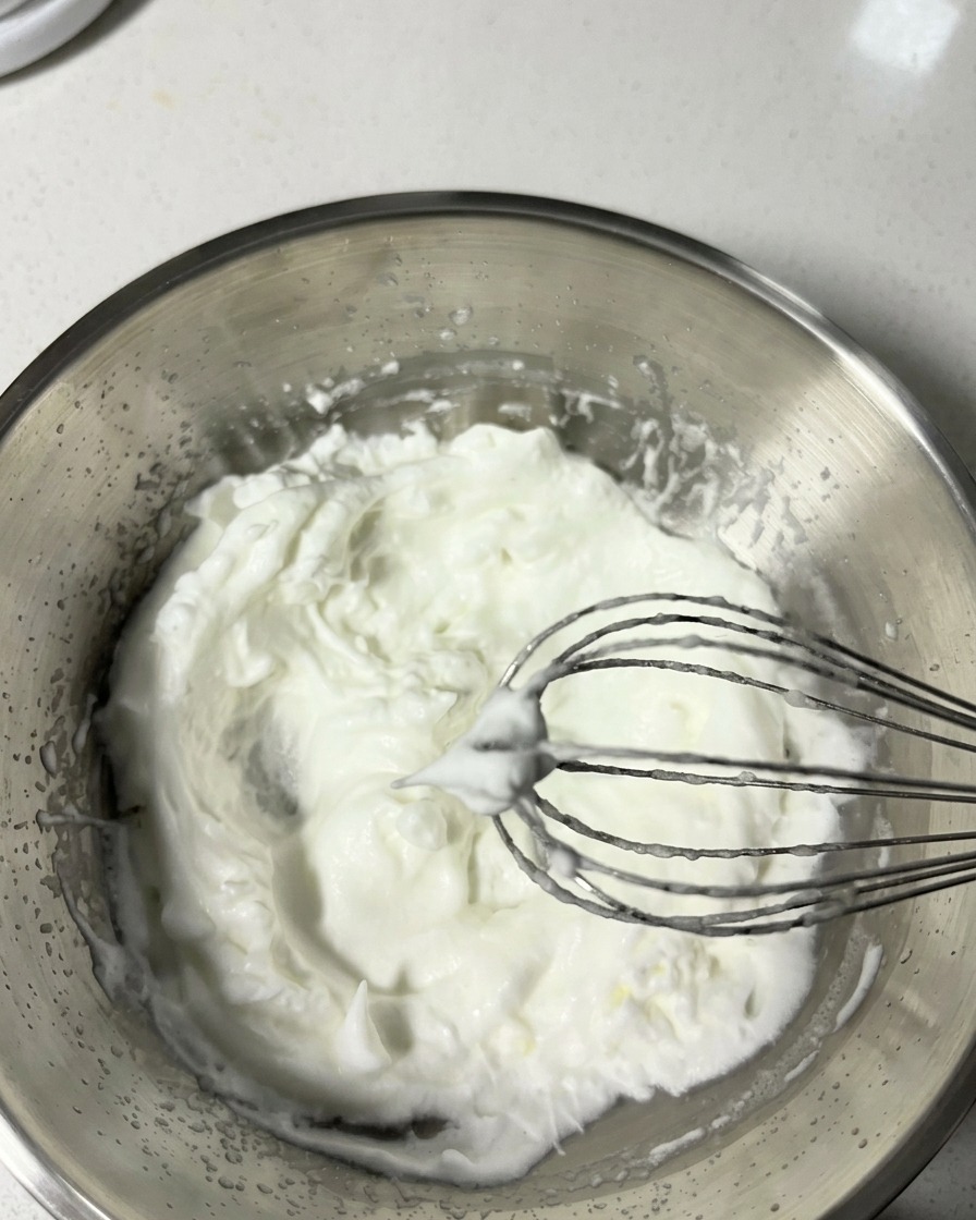 Una frusta in metallo solleva degli albumi montati da una ciotola, mostrando morbidi picchi perfetti.