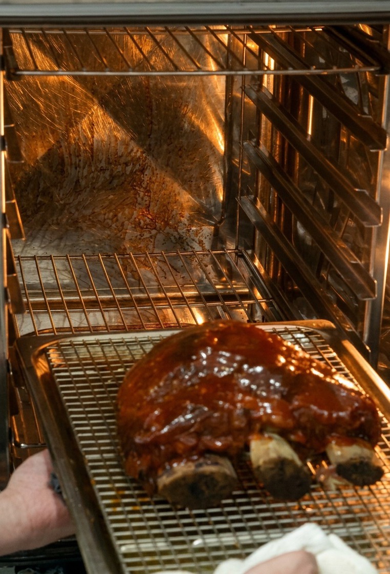 Delle mani inseriscono una teglia con le costine glassate in un forno molto caldo e illuminato.