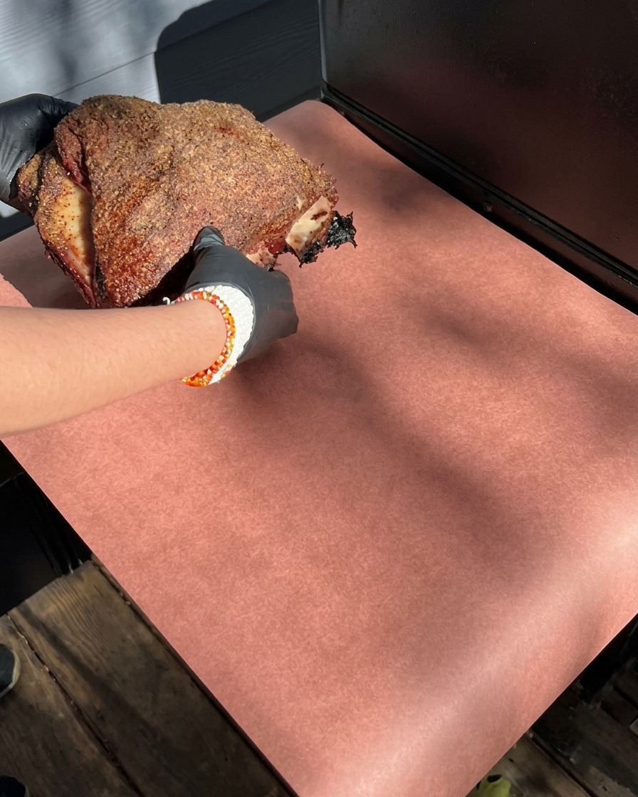 Mani guantate tengono un pezzo di costine affumicate con una crosta scura di spezie, appoggiandolo su un foglio di carta da macellaio rosa.