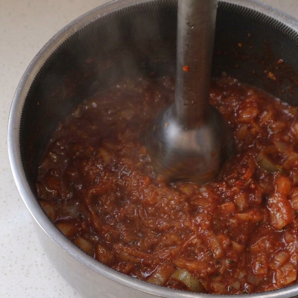 Uso di un frullatore a immersione per rendere liscia una salsa BBQ rossa in un pentolino.