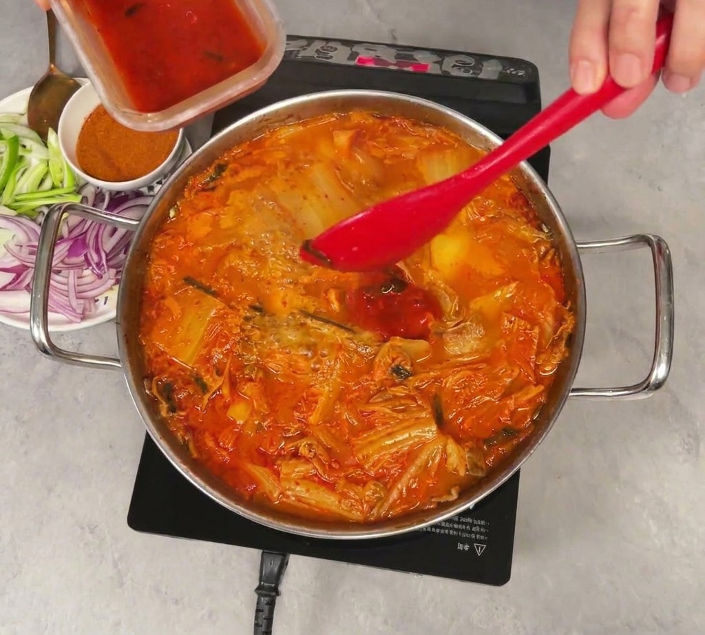 Versare il succo rosso brillante del kimchi da un piccolo contenitore di plastica in una pentola fumante di stufato, mescolato con una spatola rossa.