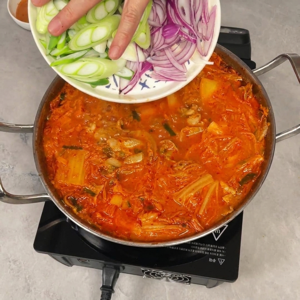 Una mano fa scivolare delle fette di cipollotto e cipolla rossa da un piatto in una pentola sobbollente di stufato di kimchi.