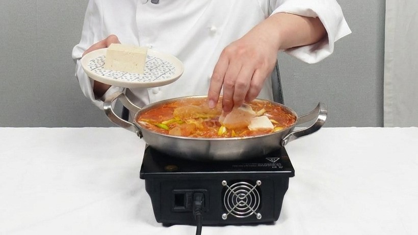 Uno chef posiziona delicatamente fette quadrate di tofu bianco in una pentola di stufato di kimchi rosso.
