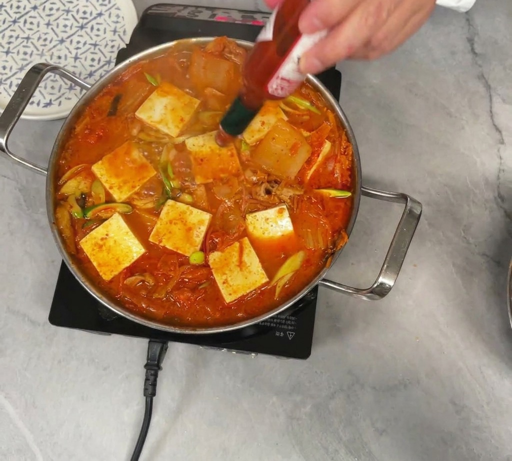 Una persona versa qualche goccia di salsa piccante Tabasco in una pentola bollente di stufato di kimchi guarnito con pezzi di tofu bianco.