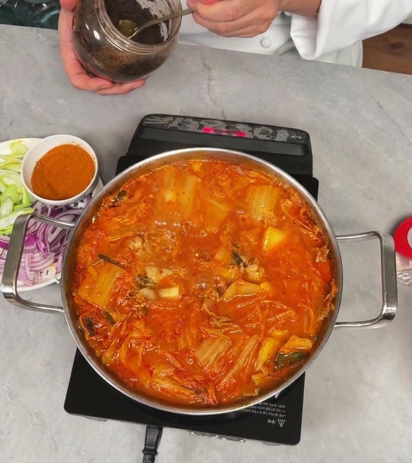 Uno chef preleva una cucchiaiata di densa pasta di soia scura da un barattolo per aggiungerla allo stufato di kimchi.