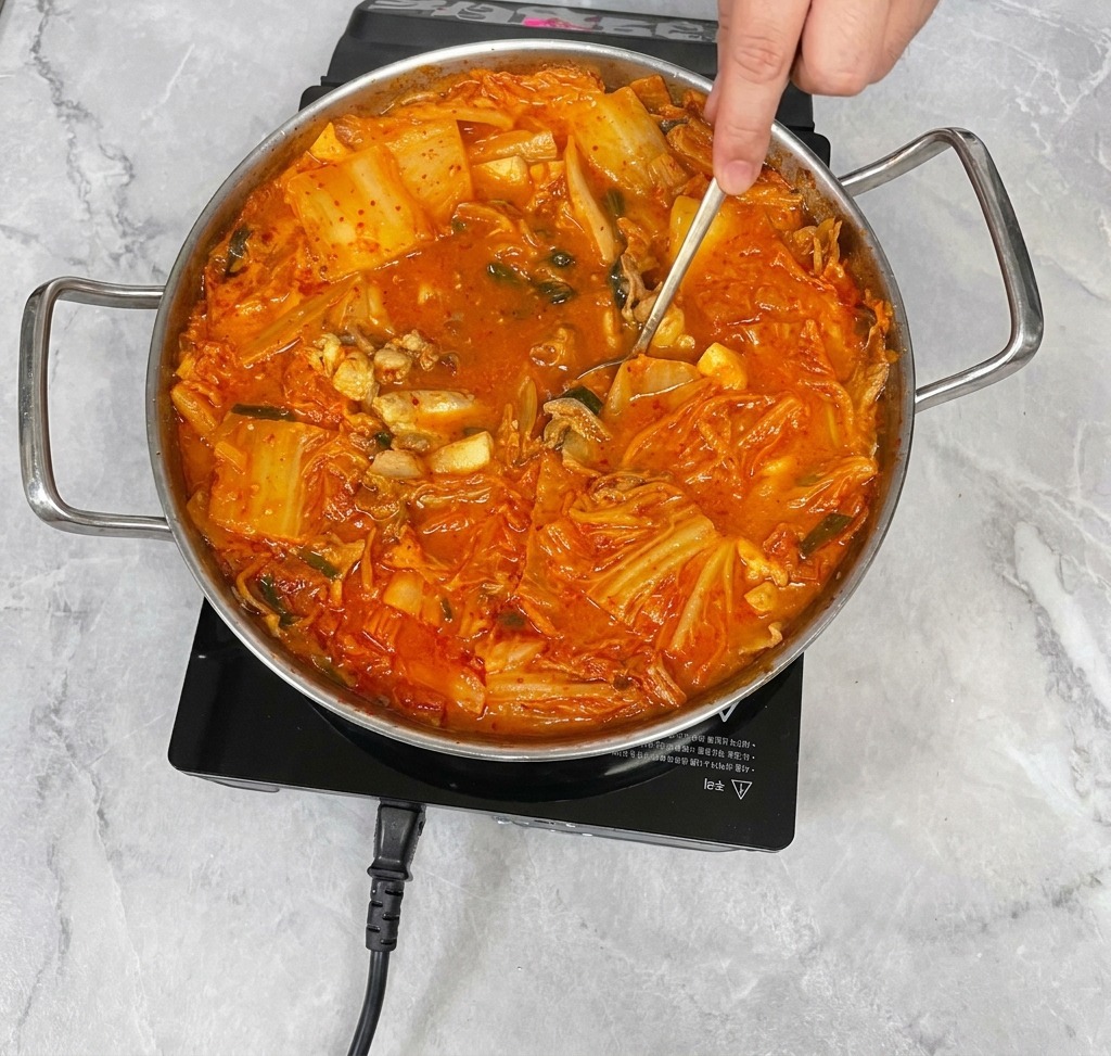 Una mano usa un cucchiaio in acciaio inossidabile per mescolare delicatamente un vivace stufato di kimchi, amalgamando i condimenti nel brodo.