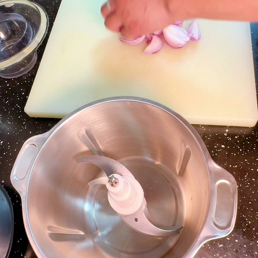 Una mano posiziona pezzi di cipolla rossa accanto a un tritatutto su un tagliere bianco.