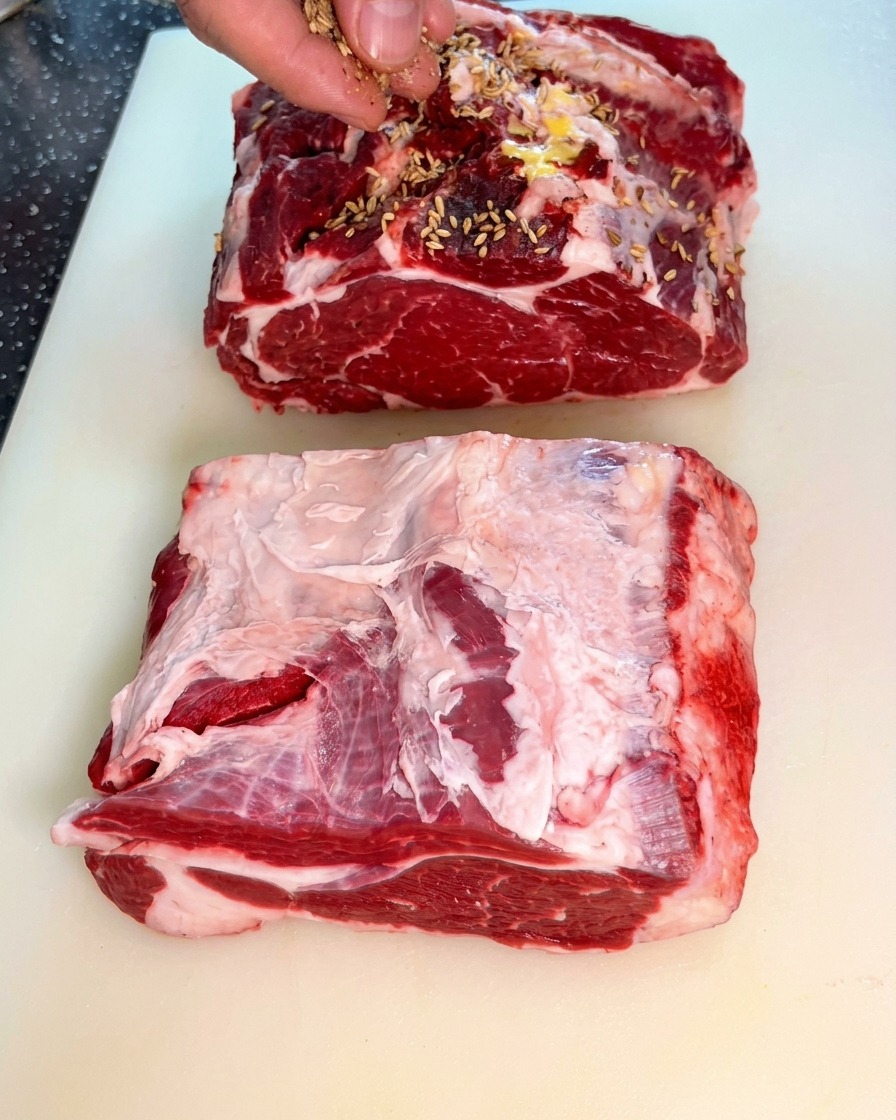 Una mano cosparge semi di finocchio su due grandi fette di reale di manzo crudo.