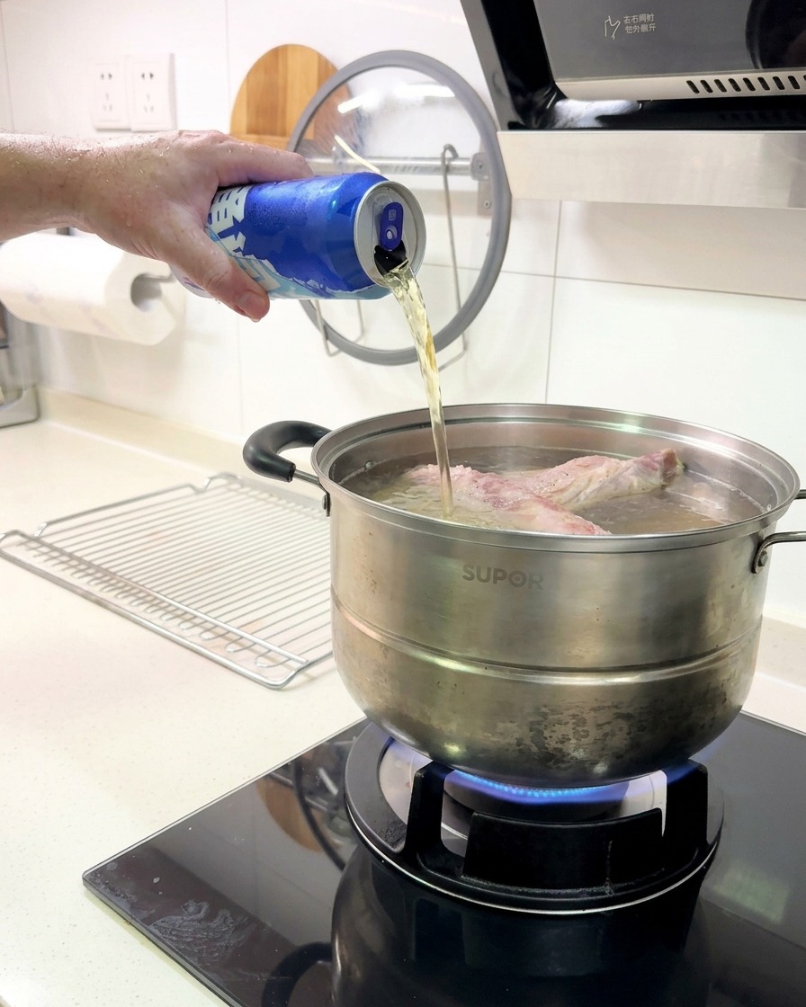 Birra versata da una lattina di alluminio blu in una pentola contenente un rack di costine che bolle su un fornello a gas.