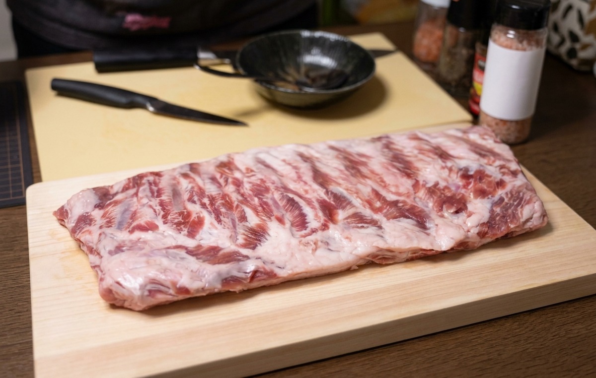 Un intero rack di costine di maiale crudo su un tagliere di legno con un coltello accanto.
