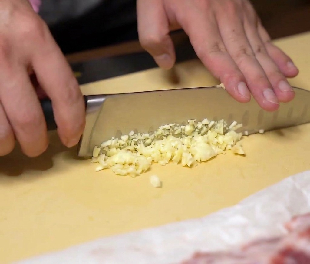 Mani che tritano finemente spicchi daglio fresco con un coltello da chef su un tagliere giallo.