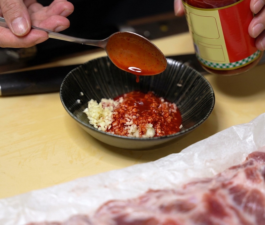 Un cucchiaio di liquido rosso di tofu fermentato aggiunto a una ciotola nera con aglio tritato.