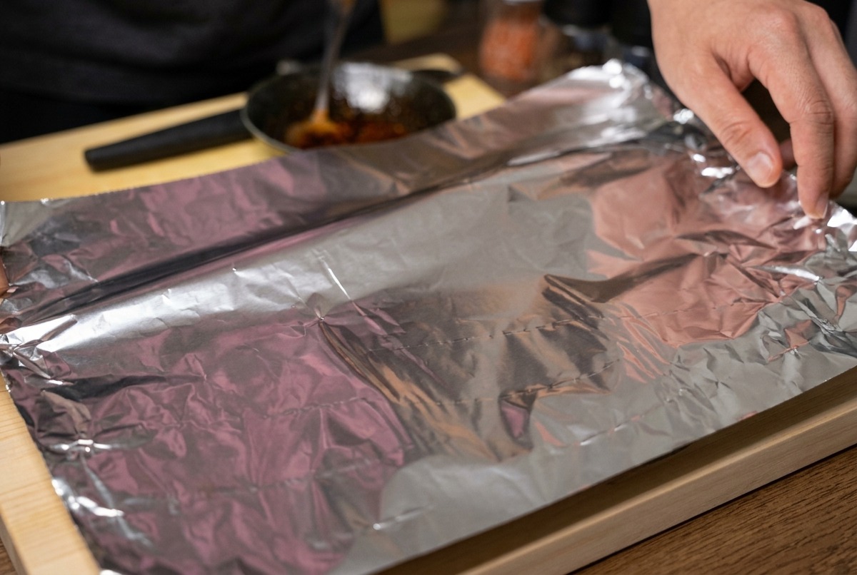 Mani che piegano un grande foglio di alluminio per avvolgere strettamente le costine marinate su un tagliere.