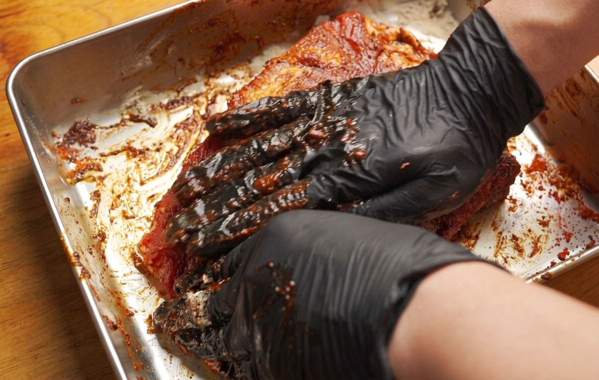 Mani guantate che massaggiano una pasta scura sulle costine di maiale.