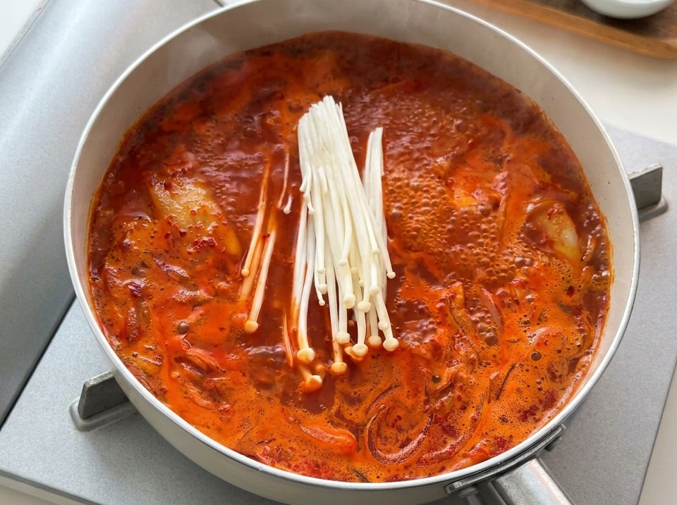 Un mazzetto di funghi enoki freschi bianchi adagiati in una padella con stufato di kimchi rosso in ebollizione.