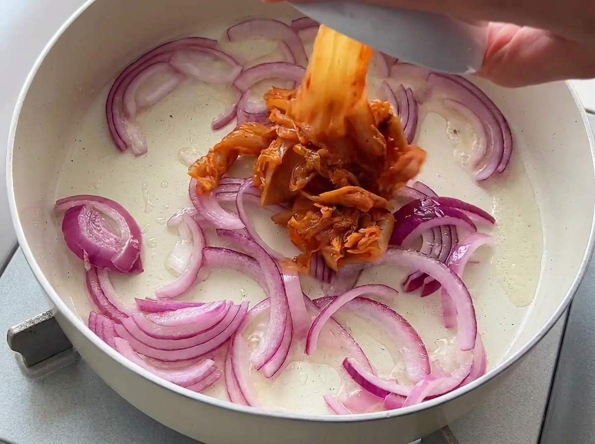 Kimchi tritato versato da una ciotolina in una padella con cipolle rosse soffritte.