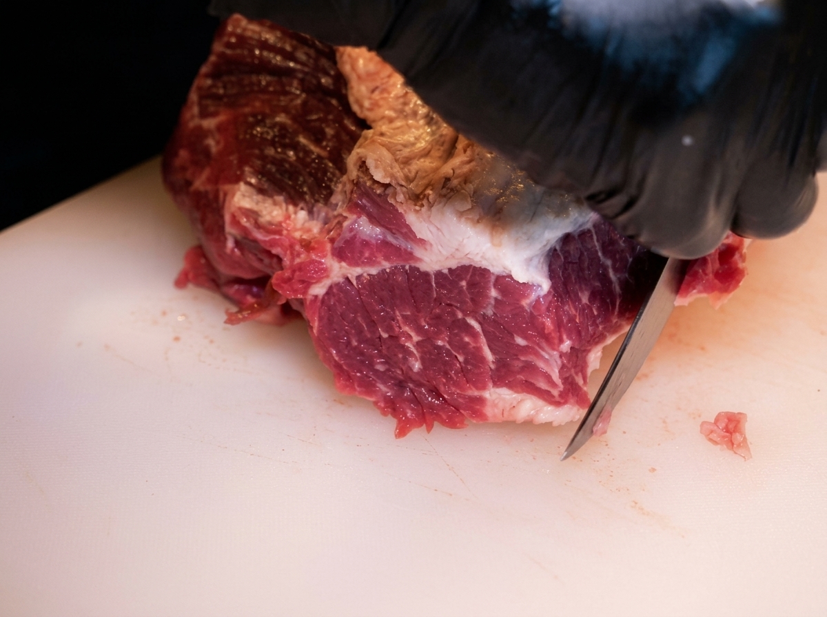 Primo piano di mani con guanti che usano un coltello per rimuovere il grasso in eccesso da una punta di petto di manzo cruda su un tagliere bianco