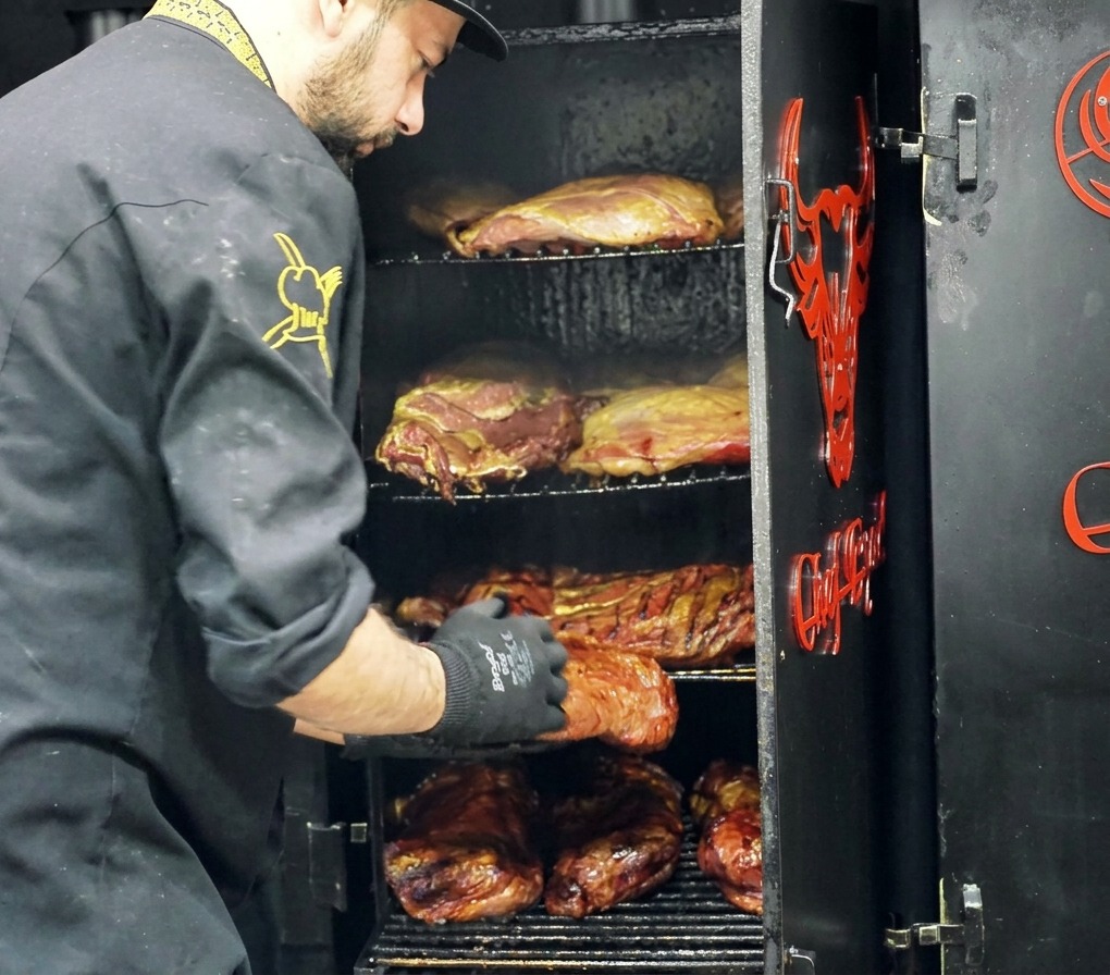 Uno chef che estrae un grosso pezzo di manzo affumicato con una crosta scura da un affumicatore multi-griglia.