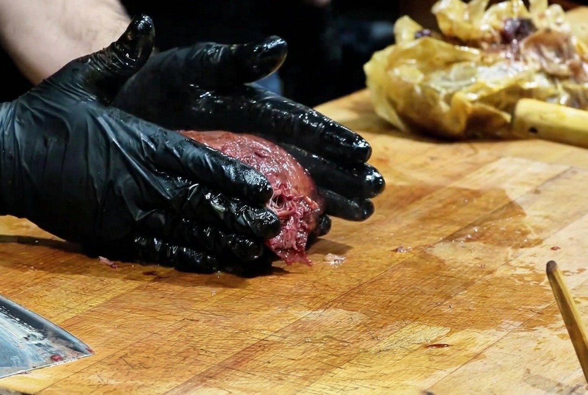 Uno chef con guanti neri che preme su un grosso brisket affumicato con crosta scura per sfilacciare la carne.