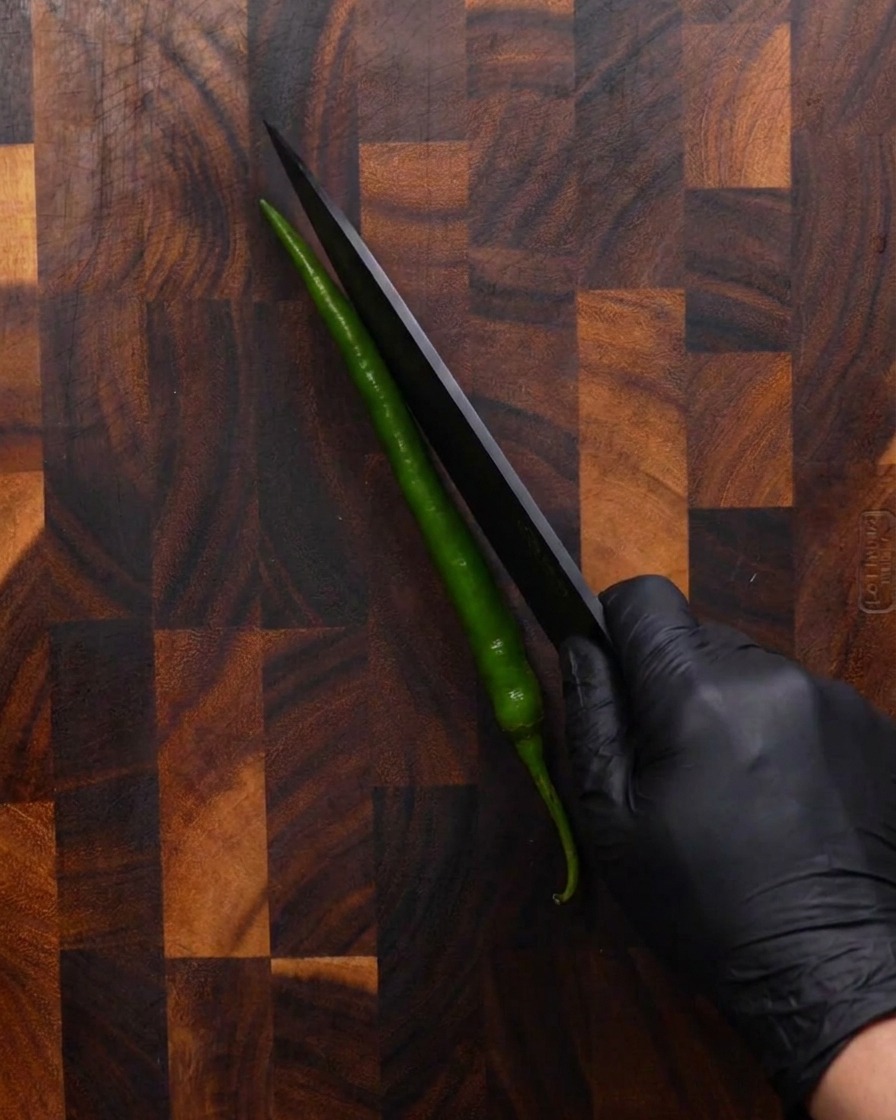 Una mano con un guanto nero usa un coltello nero affilato per tagliare in diagonale un peperoncino verde lungo su un tagliere di legno.
