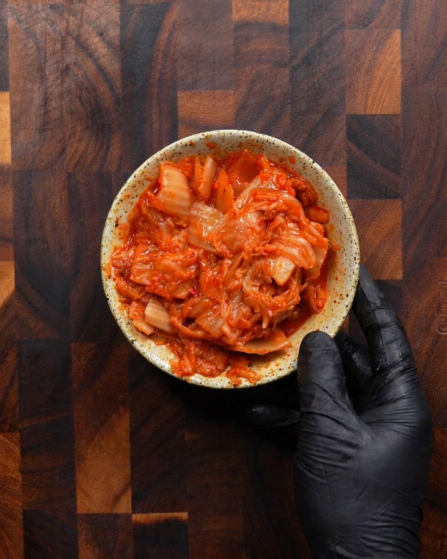 Una mano guantata tiene una ciotolina rustica colma di kimchi fermentato rosso vivo tagliato a pezzetti.