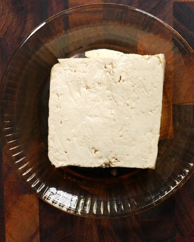 Un blocco quadrato di tofu bianco sodo in una ciotola di vetro poco profonda su una superficie di legno.