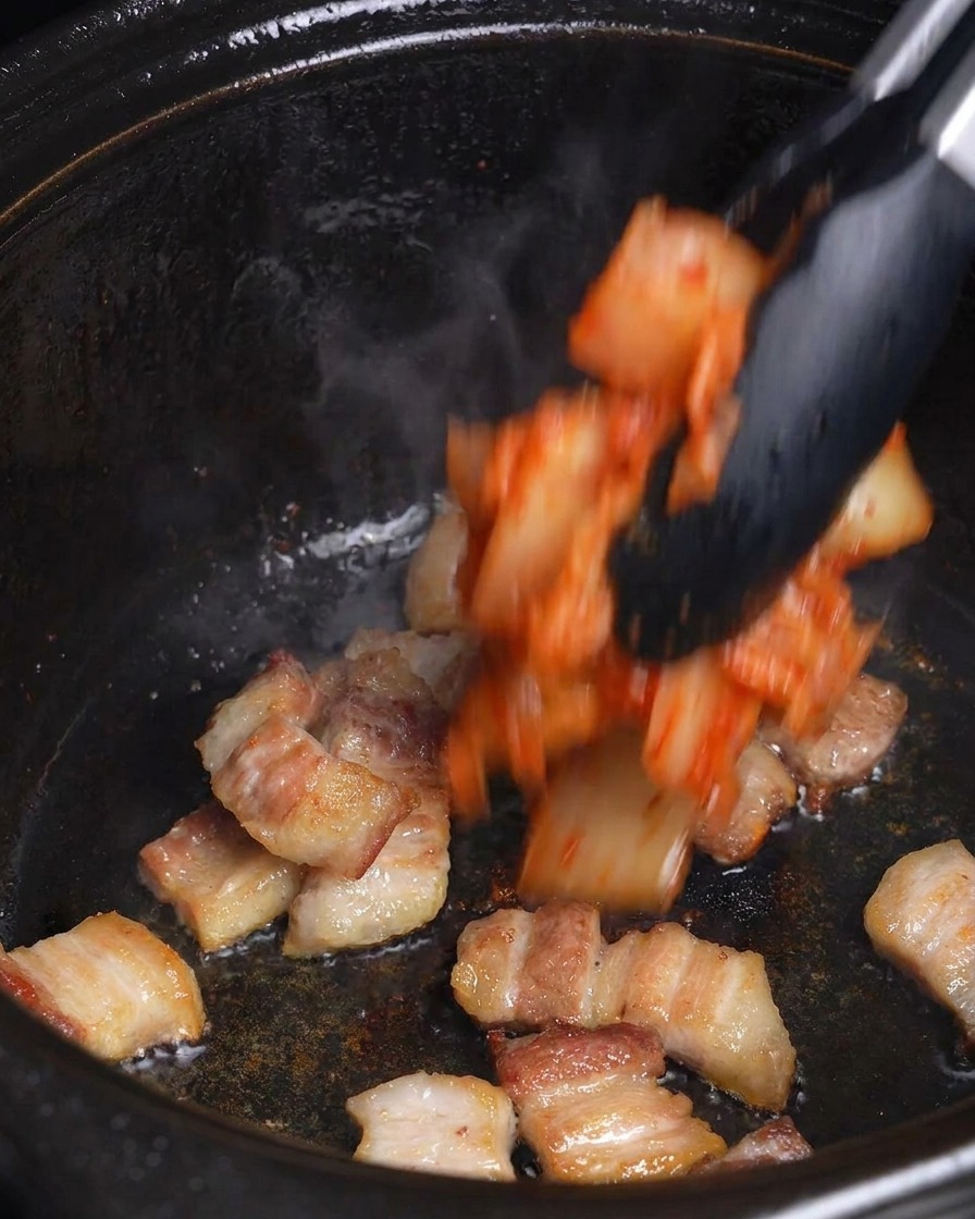 Kimchi rosso a pezzetti viene aggiunto alla pancetta rosolata in una pentola nera con delle pinze.