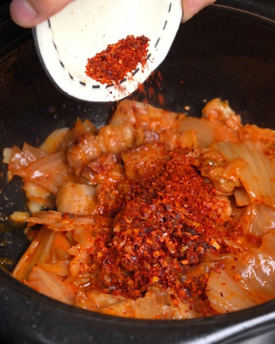 Polvere di peperoncino rosso versata da una ciotolina su kimchi e pancetta saltati in padella allinterno di una pentola nera.