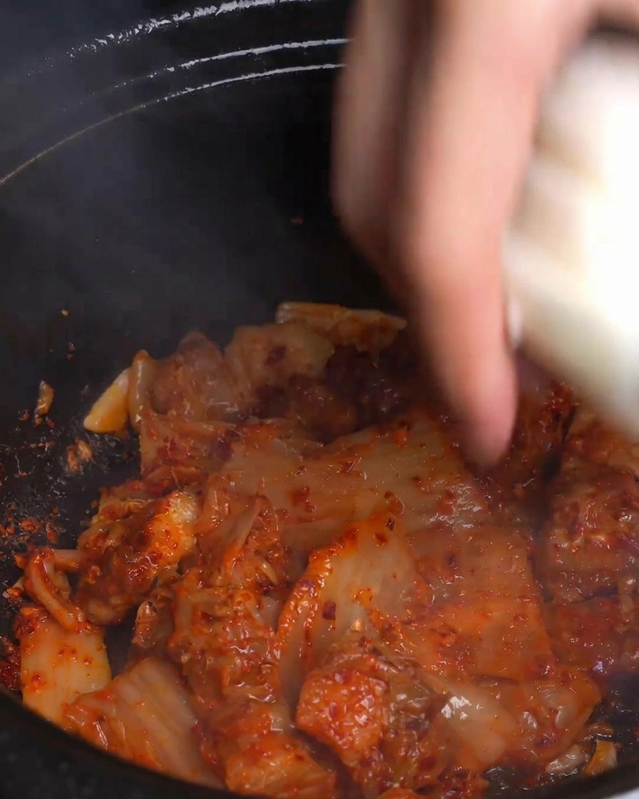 Una mano butta fette spesse di cipolla bianca fresca in una pentola calda e fumante con kimchi rosso vivo e pancetta di maiale.