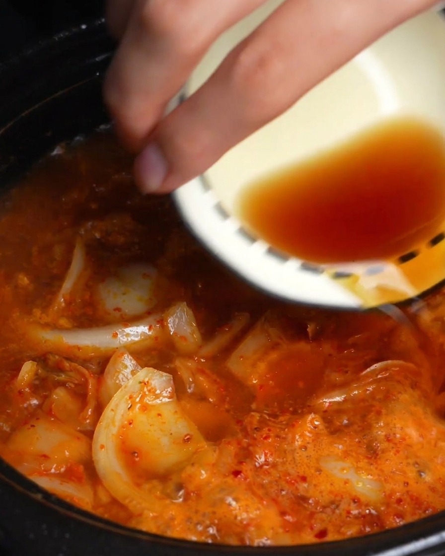 Una mano versa salsa di soia scura e salsa di pesce da una ciotolina in un brodo di kimchi rosso che sobbolle.