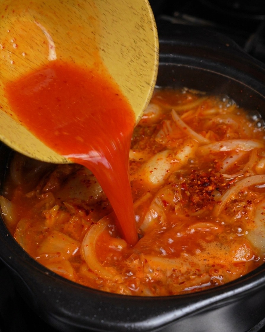 Versare succo di kimchi rosso brillante da una ciotola di legno in una pentola di stufato di pancetta e kimchi che bolle.