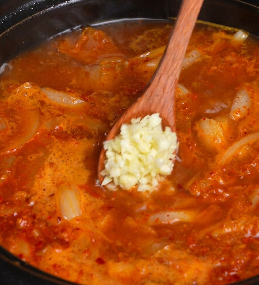 Un cucchiaio di legno aggiunge una pallina di aglio fresco tritato in una pentola fumante di zuppa di kimchi rossa.