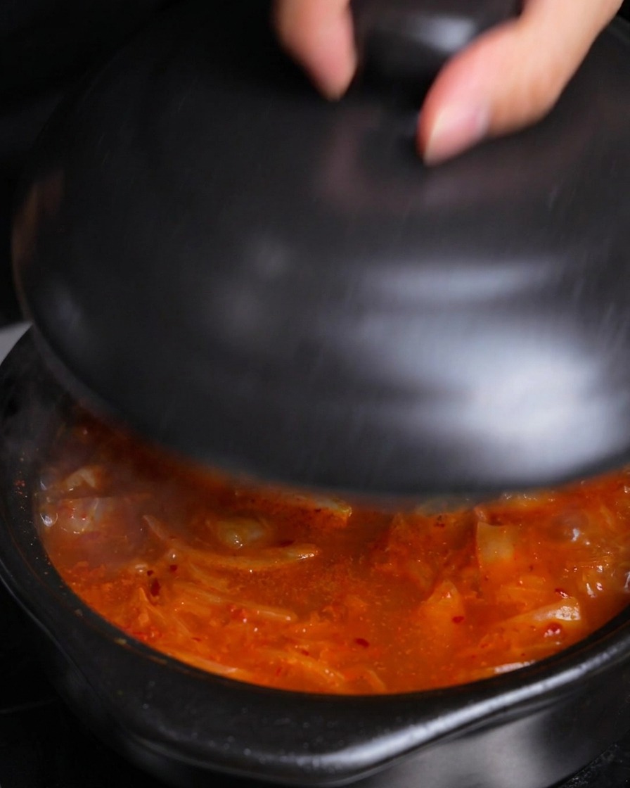 Una mano posiziona un coperchio di ceramica nera su una pentola di stufato di kimchi caldo e scoppiettante per farlo sobbollire.
