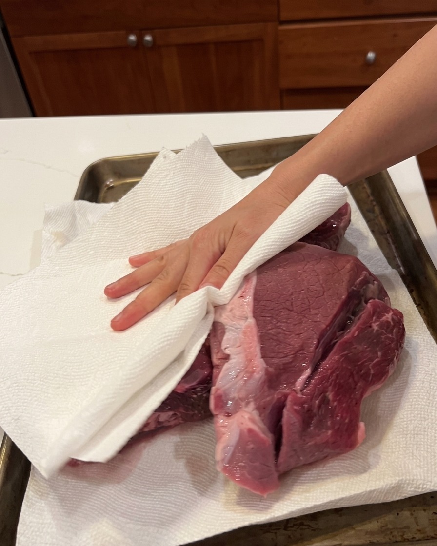 Una mano usa un tovagliolo di carta per asciugare un grosso pezzo di carne cruda su una teglia.
