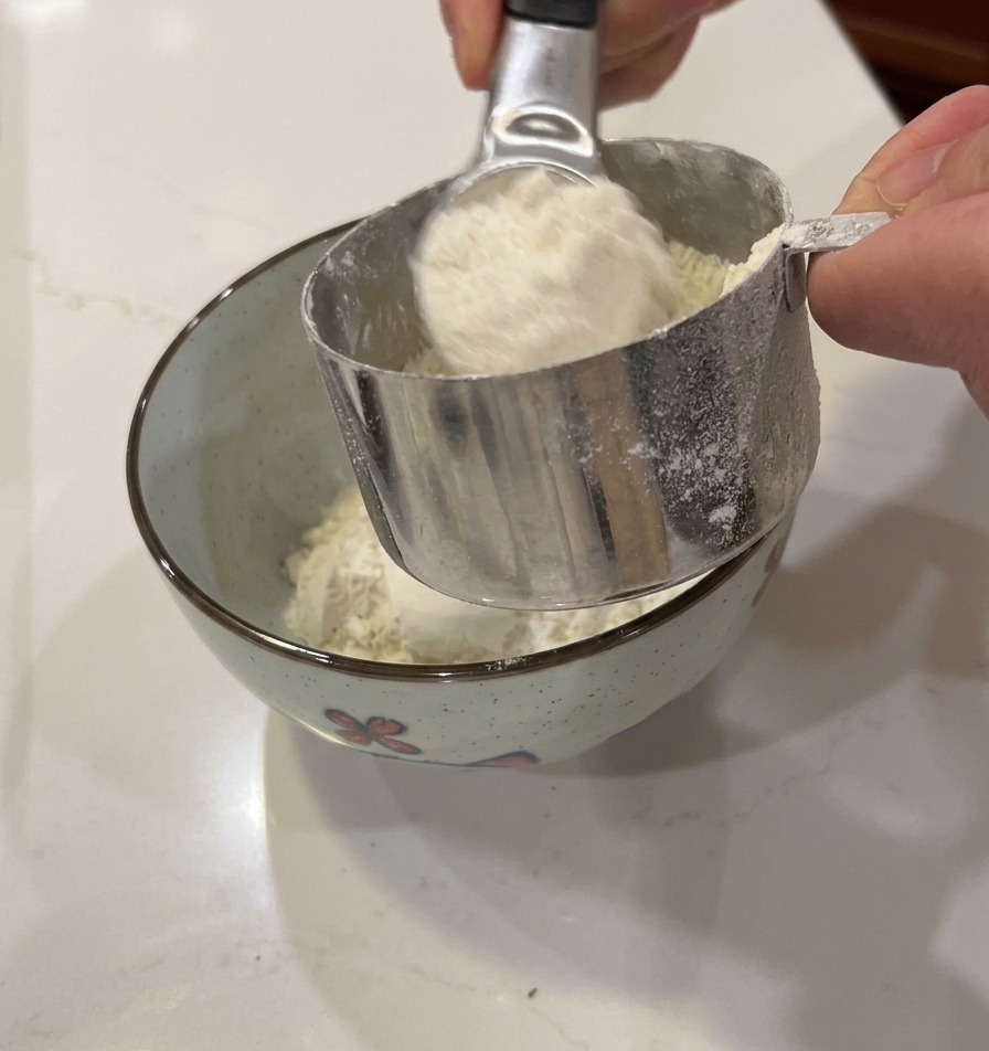Mani che versano farina bianca da un misurino in metallo in una piccola ciotola decorata.