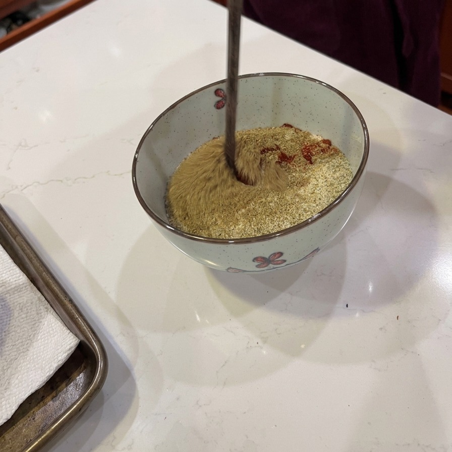 Un utensile in legno mescola un mix di farina e spezie rosso-marroni in una ciotola.