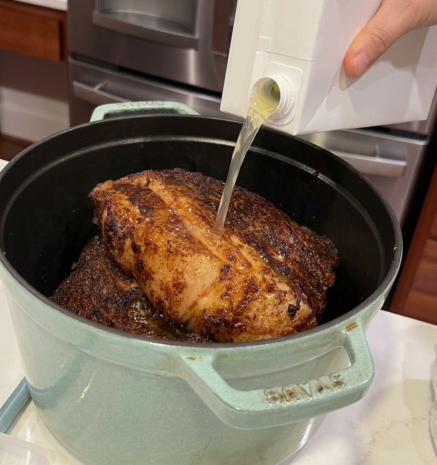 Brodo di pollo versato da un brick in una pentola di ghisa piena di grossi pezzi di manzo rosolati.