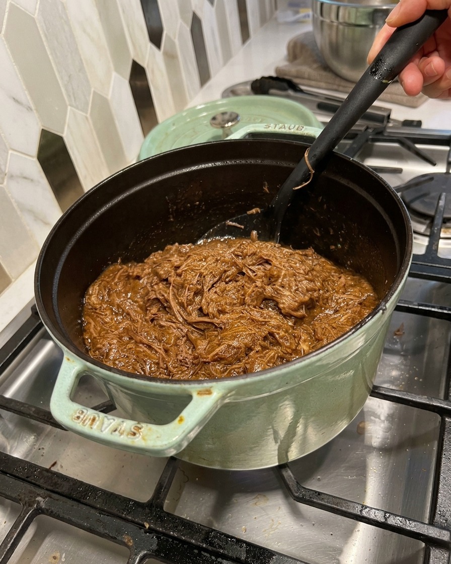Manzo sfilacciato mescolato in una ricca salsa barbecue scura in una pentola di ghisa con una spatola nera.