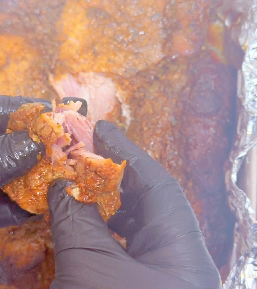 Mani con guanti neri che separano un tenero pezzo di maiale cotto lentamente, mostrando la consistenza sfilacciata e succulenta.