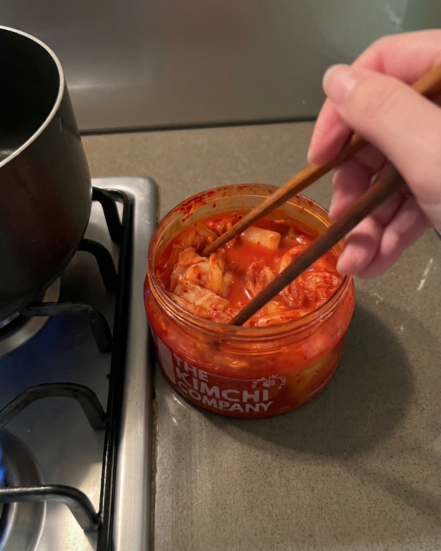 Bacchette di legno che sollevano del kimchi rosso e piccante da un barattolo di plastica trasparente.