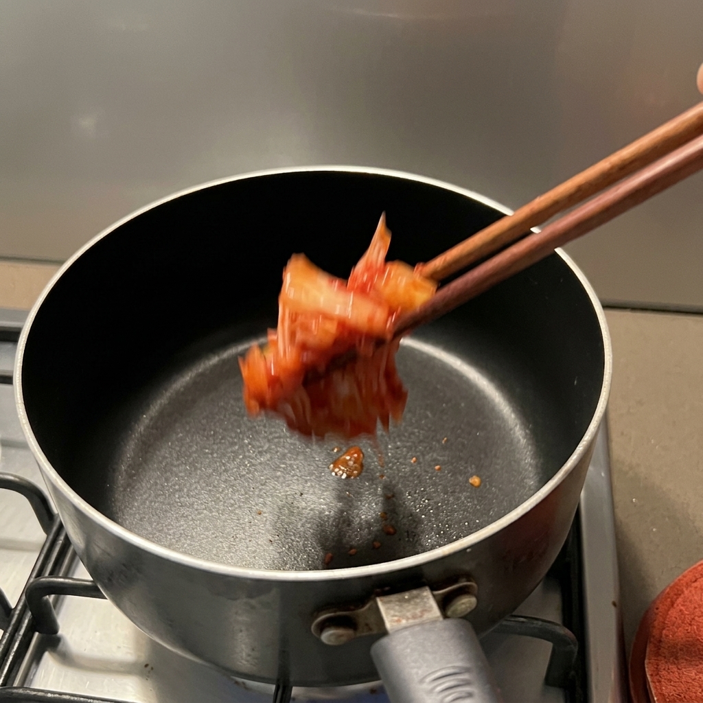 Kimchi rosso e condito che viene versato in un pentolino antiaderente usando bacchette di legno.