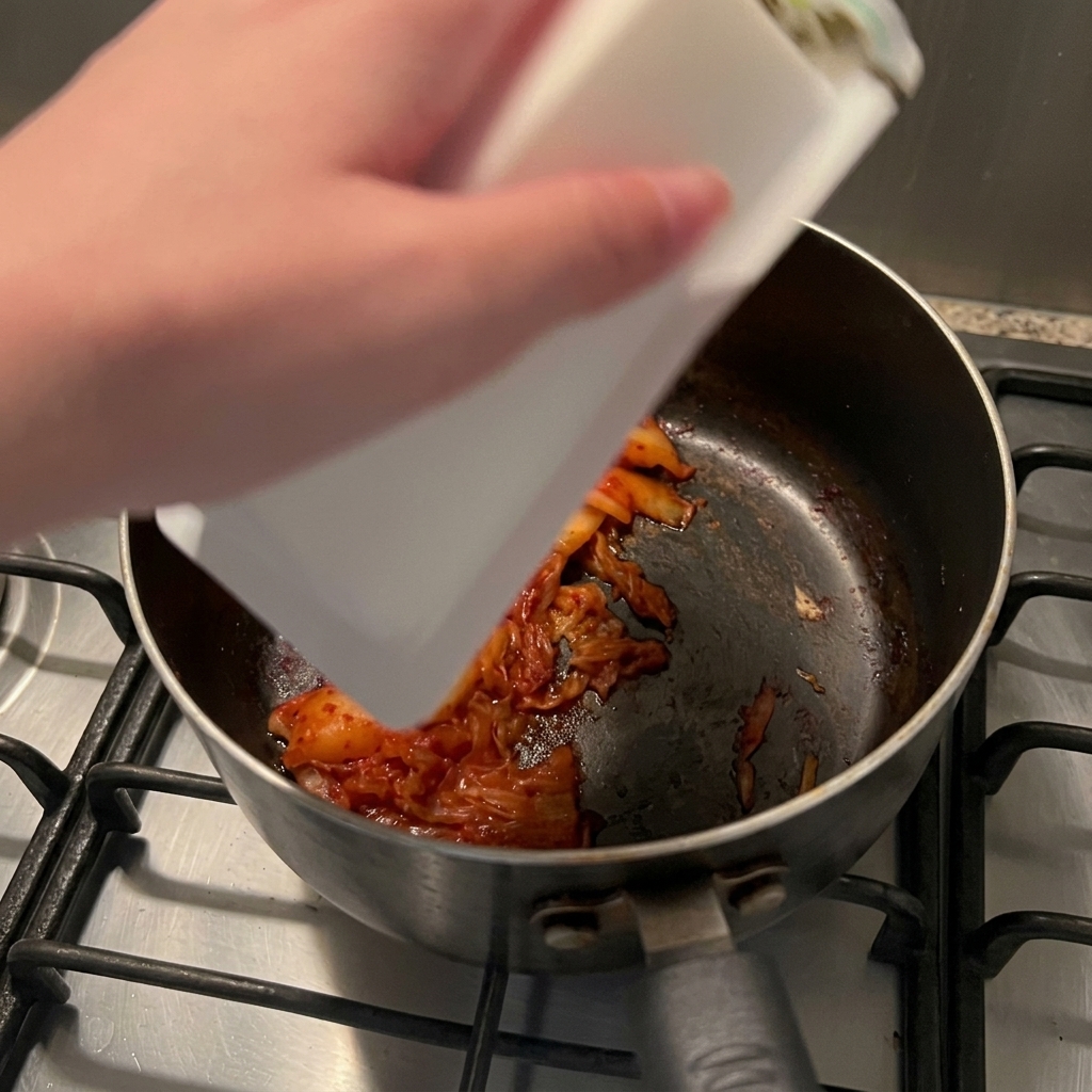 Un blocco di tofu bianco setoso che scivola dalla sua confezione in un pentolino con il kimchi soffritto.