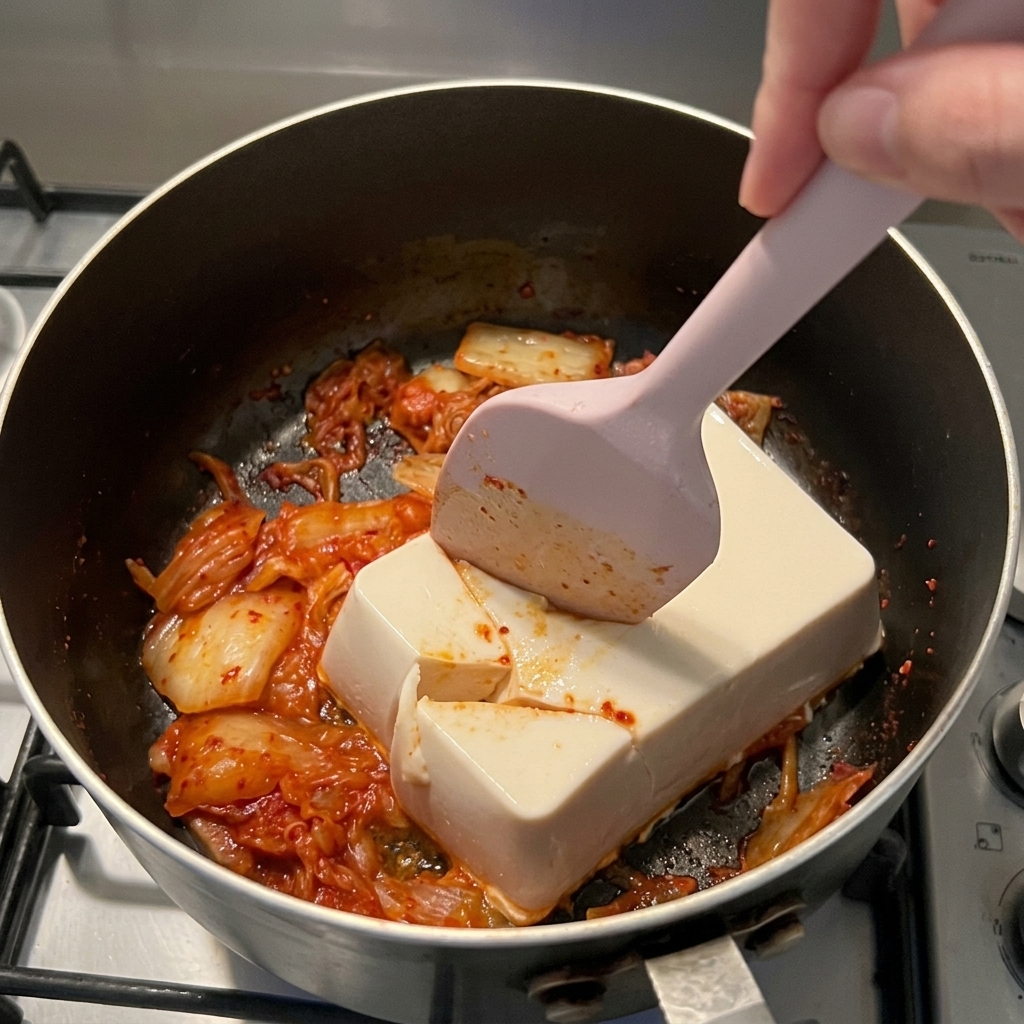 Una spatola in silicone rosa che taglia un blocco di tofu bianco sopra il kimchi rosso in un pentolino.