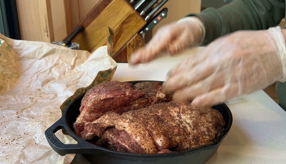 Una spalla di maiale condita adagiata in una padella di ghisa, pronta per essere infornata in un forno preriscaldato.
